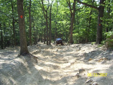 1978 Ford Bronco - A dark SUV tackles a challenging off-road trail through a wooded area on a sunny day.