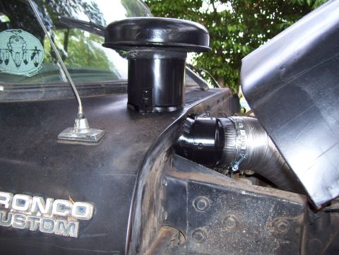 1978 Ford Bronco - Close-up of a black Ford Bronco Custom's engine bay area, showing a round black air cleaner mounted vertically and a flexible metal intake…