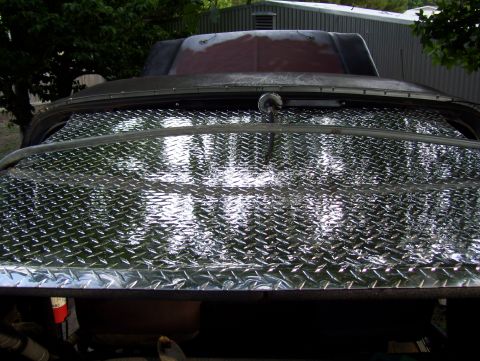 1978 Ford Bronco - Close view over the front of a truck whose hood has been skinned in polished aluminum diamond plate, reflecting the trees overhead. Beyond…