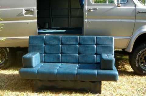 1978 Ford E250 - A blue tufted vinyl van bench sofa/bed with two loose armrest cushions sits on the grass in front of a tan van with its side cargo door sli…