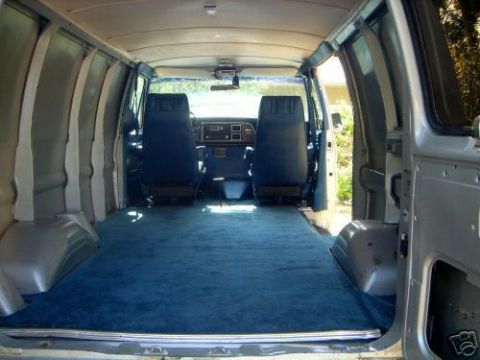 1978 Ford E250 - Interior of an empty cargo van looking forward from the open rear doors, showing blue carpeted floor and wheel wells, bare grey ribbed side…