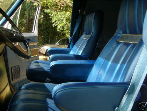 Interior view of a van showing two rows of blue bucket seats and a steering wheel. Another car is visible outside through the open door