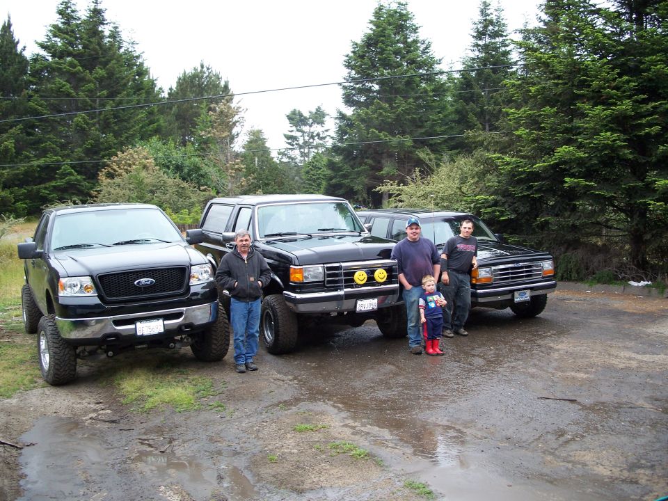 Three Ford trucks and one Ford Bronco parked on wet ground with people standing nearby