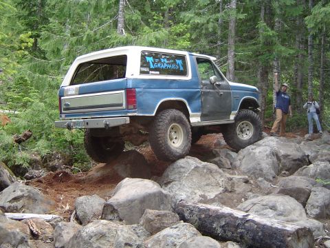 1989 Ford Bronco - A blue and white 1989 Ford Bronco tackles a challenging rocky trail, with two people observing in the background.