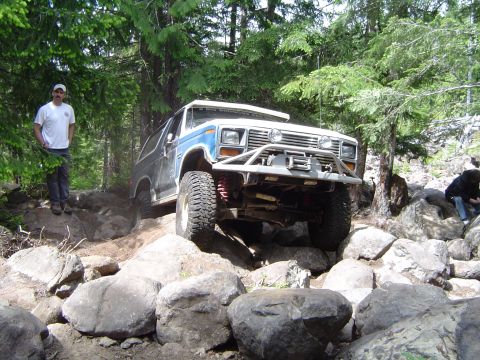 1989 Ford Bronco - A modified Ford Bronco tackles a challenging off-road course surrounded by trees and large rocks.
