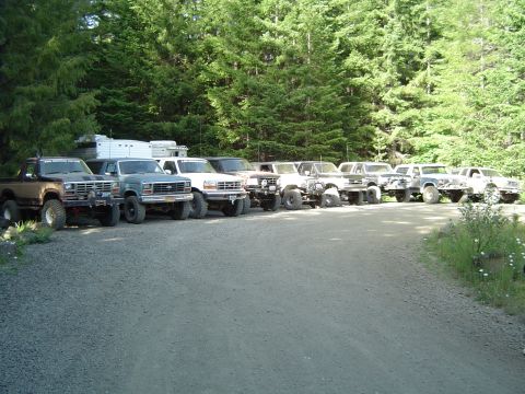 1989 Ford Bronco - A collection of off-road vehicles are lined up on a gravel road, with a dense forest behind them.