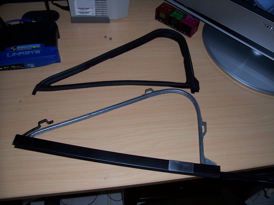 Two automotive window trim pieces, one black rubber seal and one chrome and black metal frame, lie on a wooden desk
