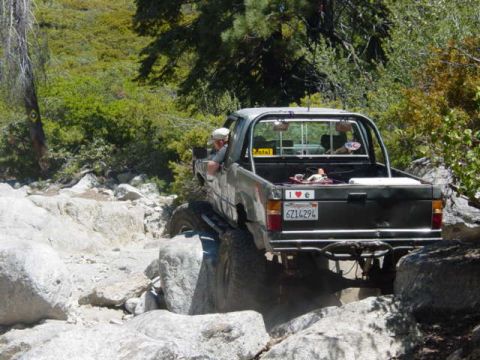 1986 Ford Bronco - A pickup truck navigates a rocky off-road trail with trees and brush in the background.