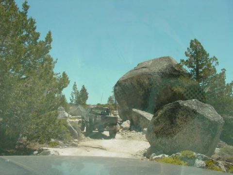 1986 Ford Bronco - A dark off-road truck navigates a boulder-strewn path under a clear blue sky.