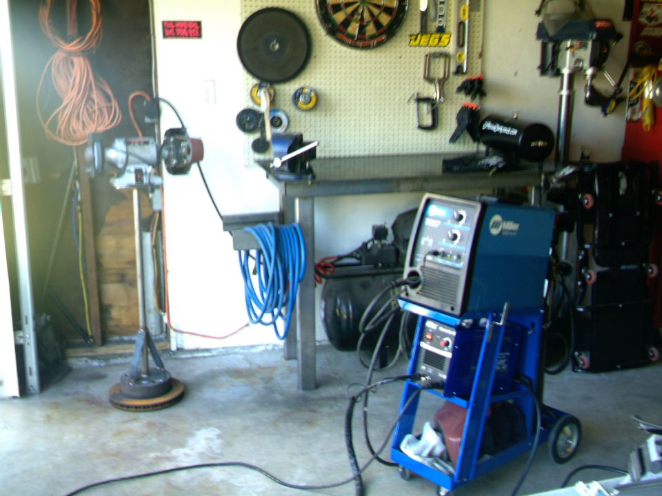 Welding machine on a blue cart, air compressor, and various tools on a pegboard wall in a garage