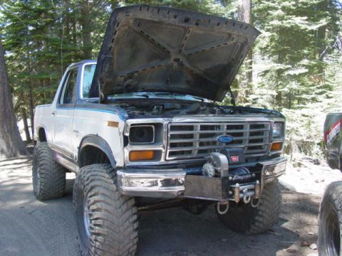 1986 Ford Bronco - A modified white Ford Bronco with its hood up sits off-road, with another vehicle partially visible to the right.