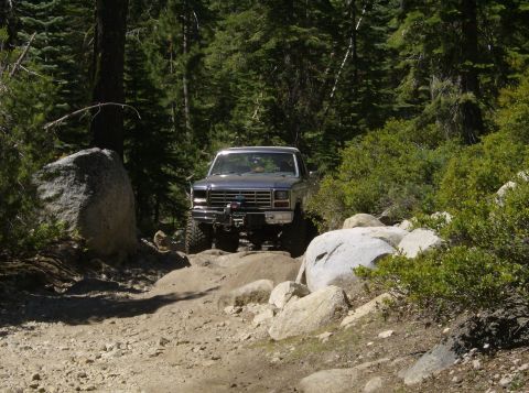 1986 Ford Bronco - A gray Ford Bronco navigates a rocky off-road trail surrounded by trees.