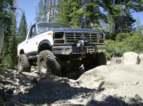 1986 Ford Bronco - Modified Ford Bronco with a winch drives over a boulder-strewn path in a wooded area.