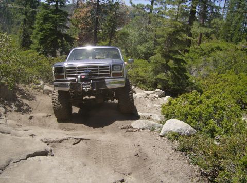 1986 Ford Bronco - A lifted Ford Bronco navigates a challenging rocky path through a wooded area.