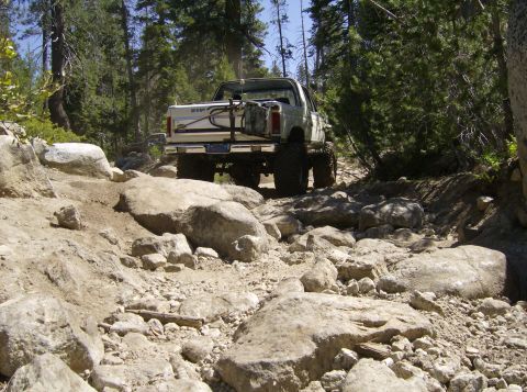 1986 Ford Bronco - This modified SUV is tackling a challenging, boulder-strewn path through a wooded area.