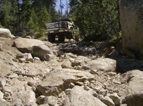 1986 Ford Bronco - A white pickup truck navigates a challenging, rocky trail surrounded by trees.