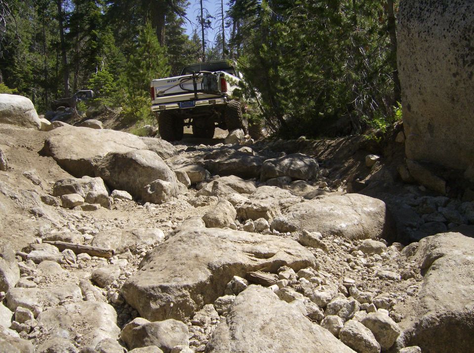 White pickup truck with spare tire rack driving over large rocks on a wooded trail