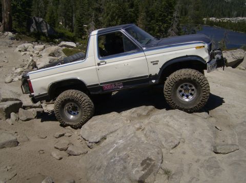1986 Ford Bronco - A white and gray Ford Bronco navigates a rocky terrain near a body of water.