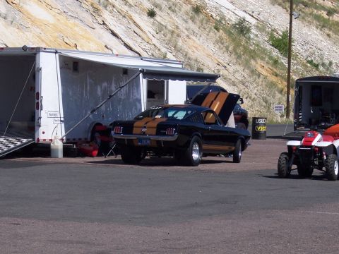 1972 Ford Ranchero - A black coupe with gold racing stripes is parked next to a trailer at an outdoor event.
