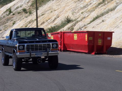 1972 Ford Ranchero - A black Ford pickup truck drives past two large red dumpsters next to a rocky hillside.