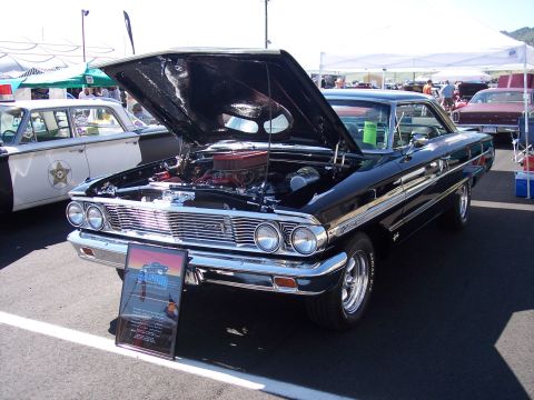 1972 Ford Ranchero - A black 1964 Ford Galaxie coupe is displayed with its hood open, revealing the engine, next to a police car.