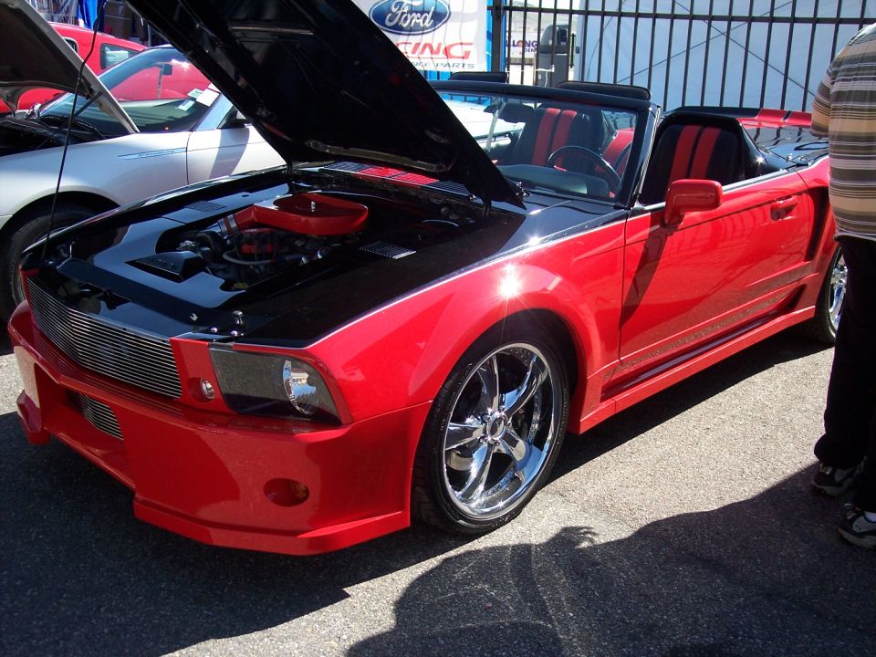 Red convertible with black hood and racing stripes, hood open showing engine. Another car is visible behind it