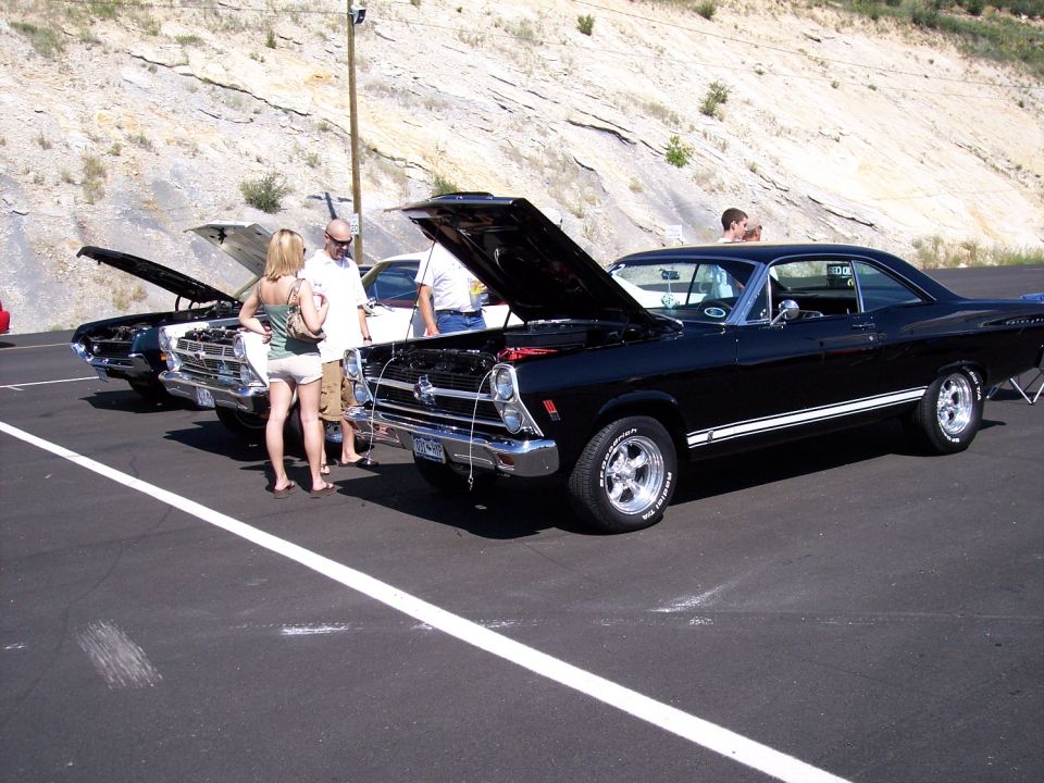 Black coupe with white racing stripes, hood open, parked next to other cars