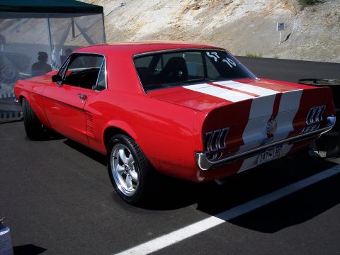 1972 Ford Ranchero - A red 1967 Ford Mustang coupe with white racing stripes is parked on asphalt, showing its rear three-quarter view.