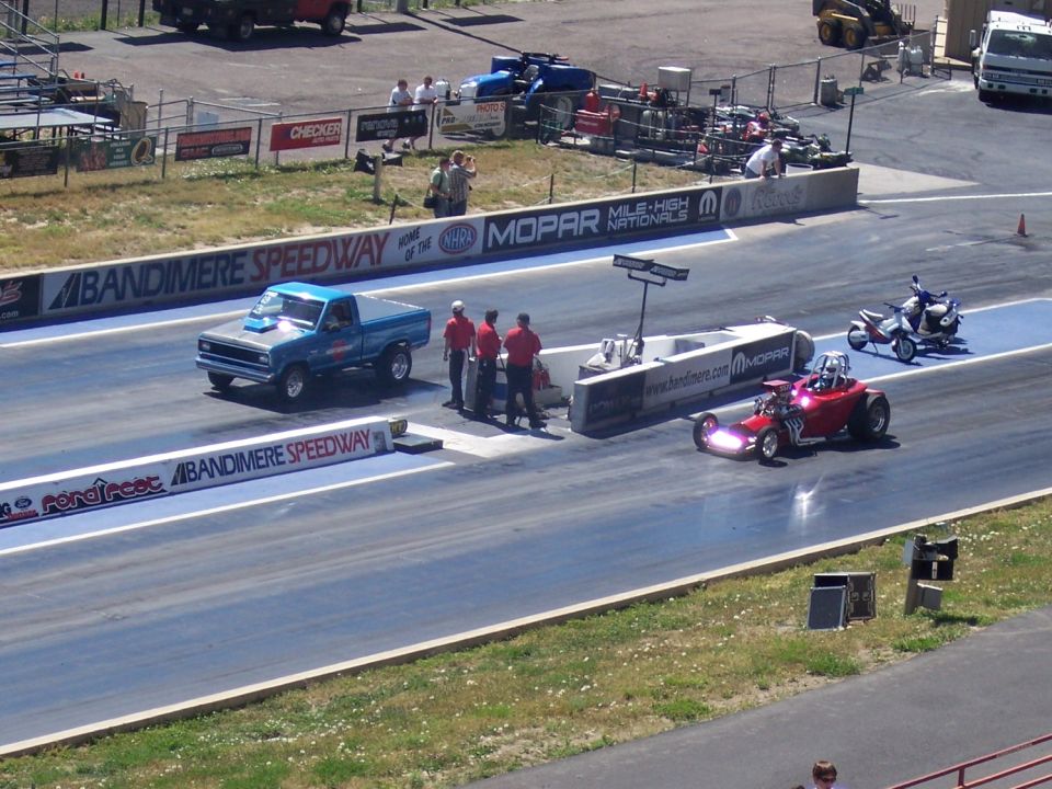 Drag strip with a blue pickup truck, a red dragster, and a scooter