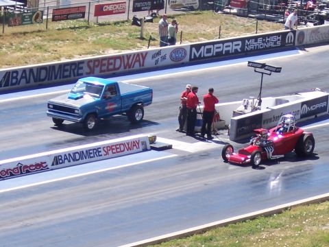 1972 Ford Ranchero - A blue pickup truck and a red dragster are on the race track at Bandimere Speedway.