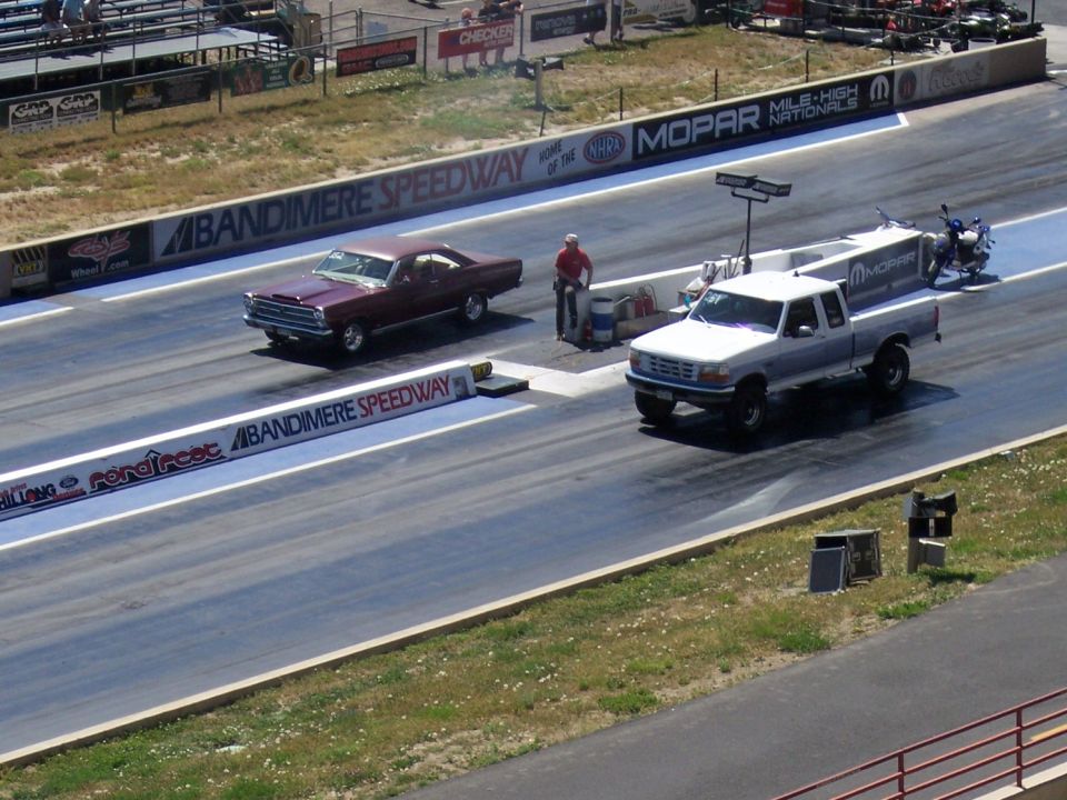 Two vehicles racing on a drag strip, one a maroon coupe and the other a white pickup truck towing a motorcycle
