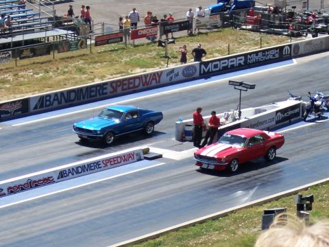 1972 Ford Ranchero - Two cars race down the drag strip at Bandimere Speedway, with spectators in the stands.