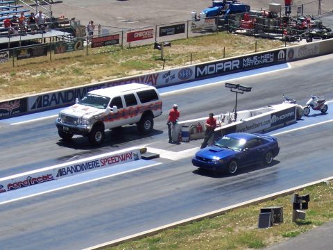1972 Ford Ranchero - Two vehicles race down the drag strip at Bandimere Speedway, with spectators in the stands.