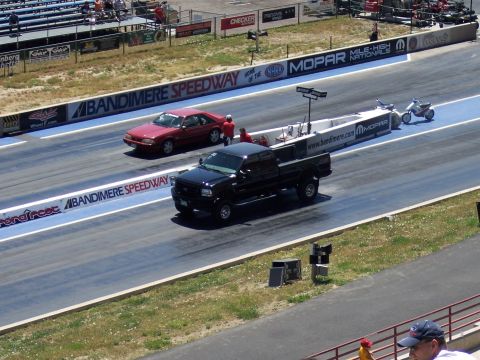 1972 Ford Ranchero - A black pickup truck and a red car are racing down a drag strip at Bandimere Speedway.