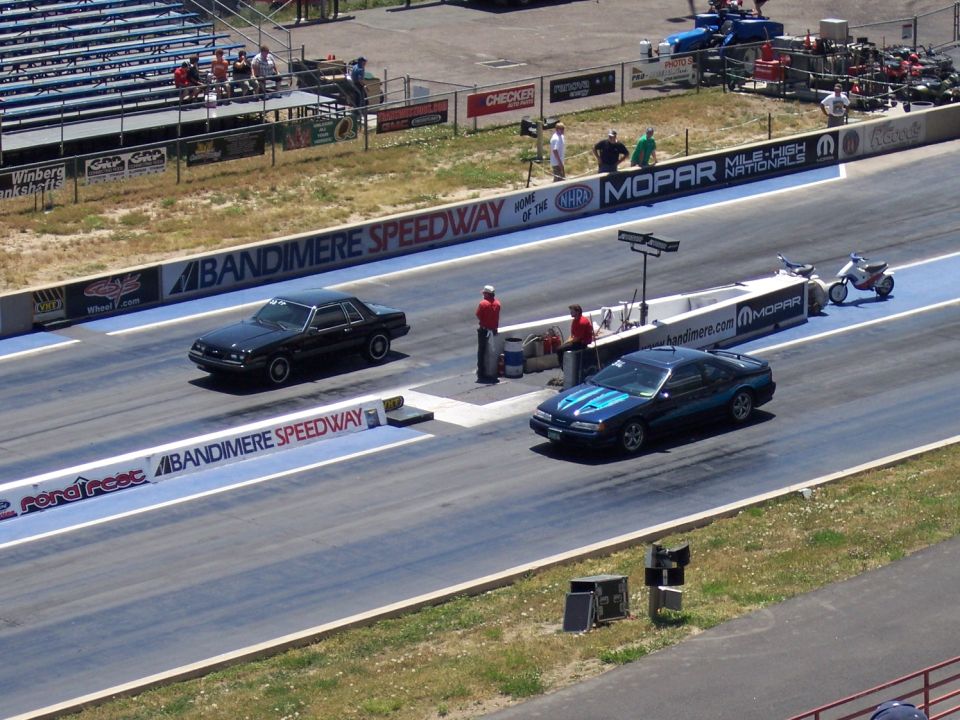 Two cars racing on a drag strip, with spectators in the stands