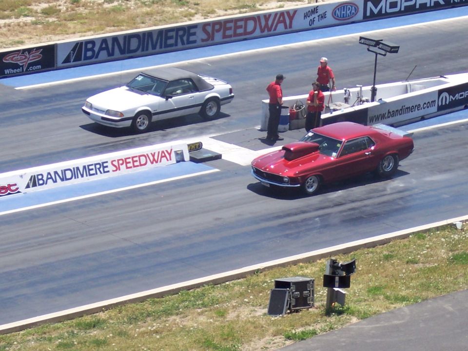 Red 1970s muscle car with hood scoop on drag strip, white convertible in background