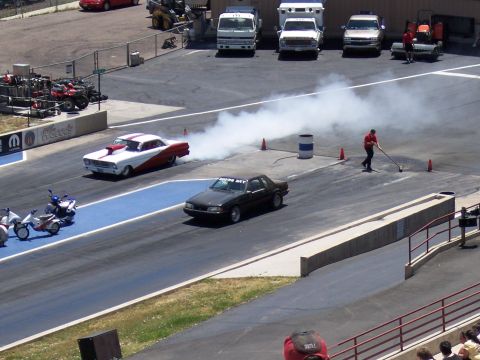 1972 Ford Ranchero - Two cars race down a drag strip, one performing a burnout as a track worker cleans the surface.