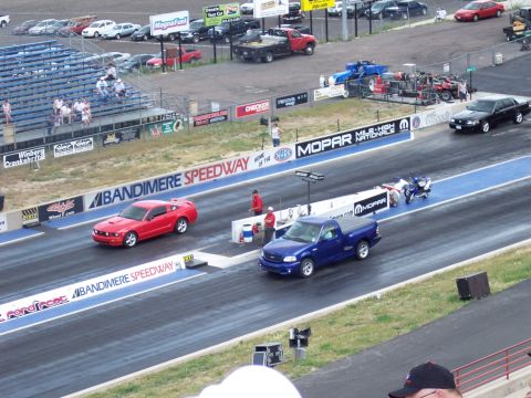 1972 Ford Ranchero - Two vehicles race down the drag strip at Bandimere Speedway, with spectators in the stands.