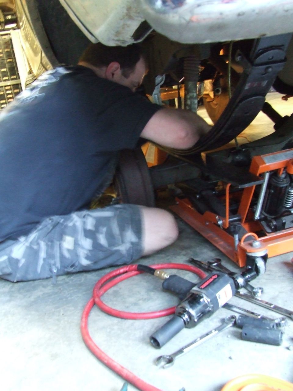Mechanic working on rear leaf spring suspension with impact wrench and air hose nearby