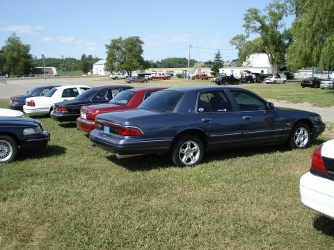2000 Ford Crown Victoria - A row of sedans are parked on a grassy field under a clear blue sky.
