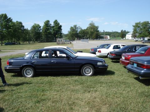 2000 Ford Crown Victoria - Several cars are parked on a grassy field under a clear blue sky.