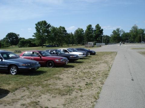 2000 Ford Crown Victoria - Multiple sedans are parked in a row on a grassy area under a clear blue sky.