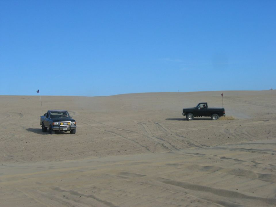 Two pickup trucks driving on sand dunes under a clear blue sky
