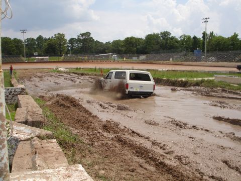 1989 Ford Bronco - A white 1989 Ford Bronco navigates a muddy track at a speedway, kicking up dirt and water.