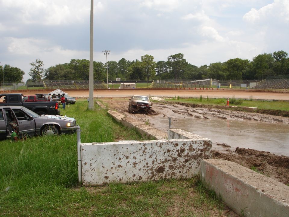 Truck stuck in mud pit at a speedway, other cars parked nearby
