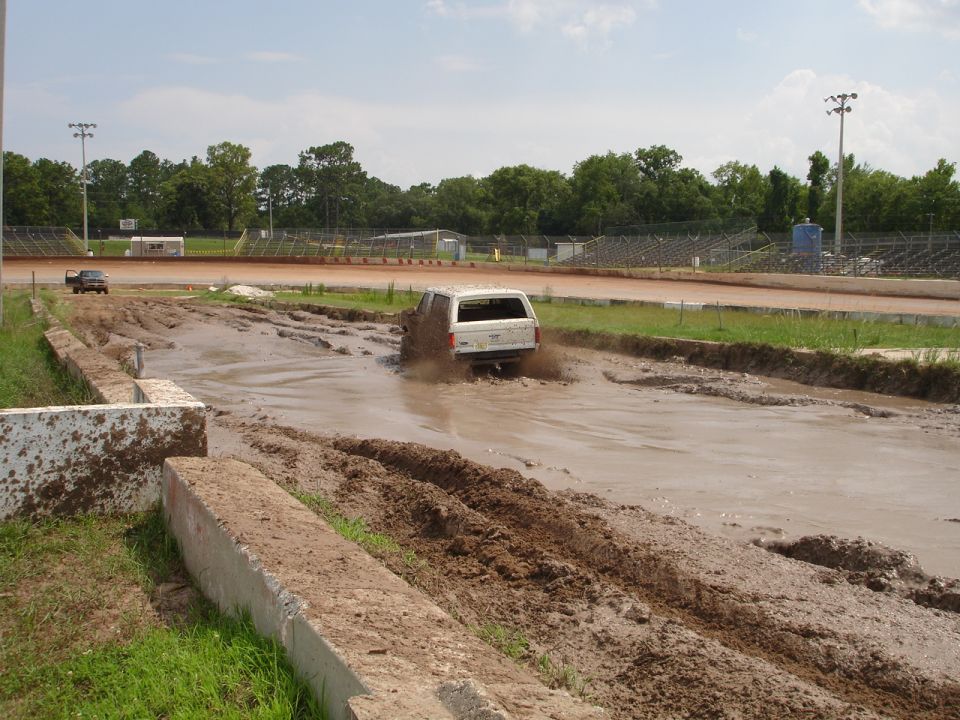 White SUV splashing through a mud pit at a race track. Another truck is visible in the background
