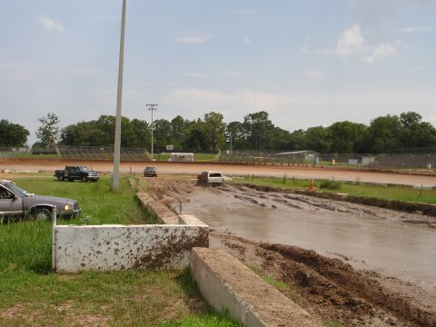 1989 Ford Bronco - Vehicles navigate a muddy track at a speedway, with spectators' seating visible in the background.