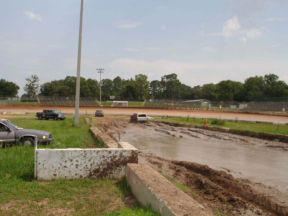 Mud bog track with a few vehicles, including a white SUV stuck in the mud. A dark pickup truck and a gray car are parked nearby