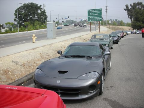 1998 Ford Crown Victoria - A line of cars, including a gray Dodge Viper, are parked along a roadside with traffic in the background.