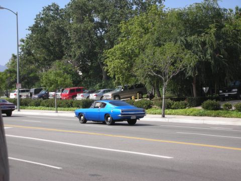 1998 Ford Crown Victoria - A blue 1967 Plymouth Barracuda coupe is seen driving on a street with other vehicles parked in the background.